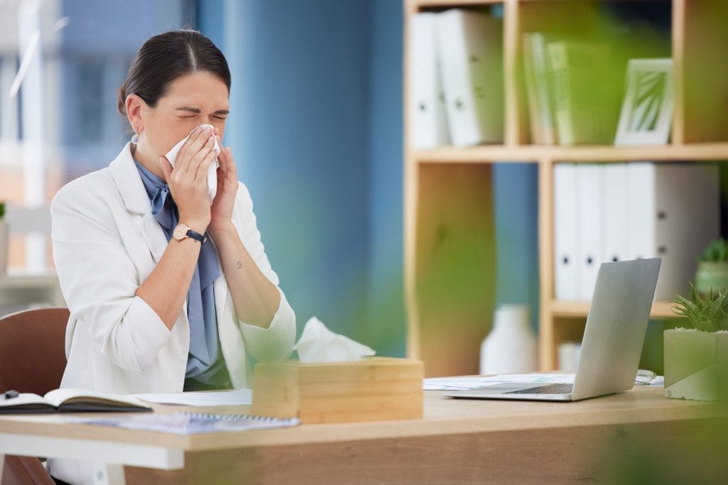 Woman Sneezing at Office Desk Indicating Need for Moisture Air Testing Service